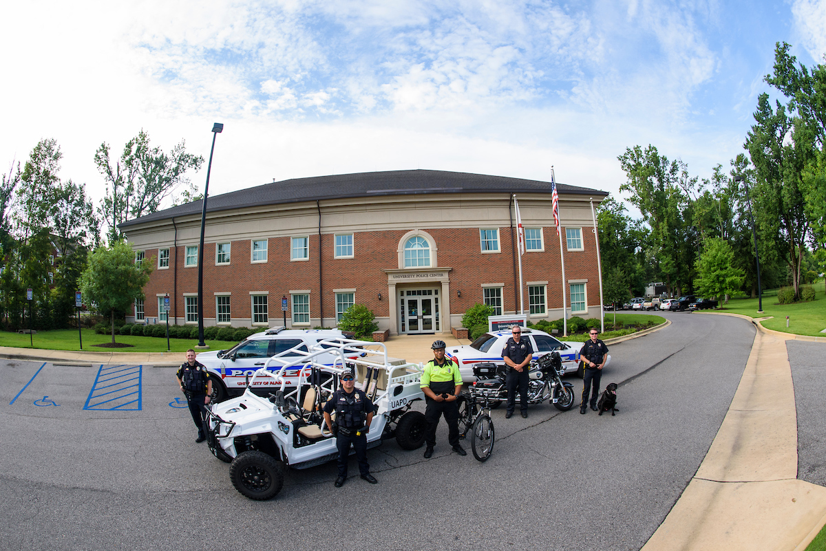 University police officers standing in front of the UA Police Center with patrol cars, bicycles and a police dog.