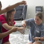Three female university students work to untangle a spool of lights in a dorm room