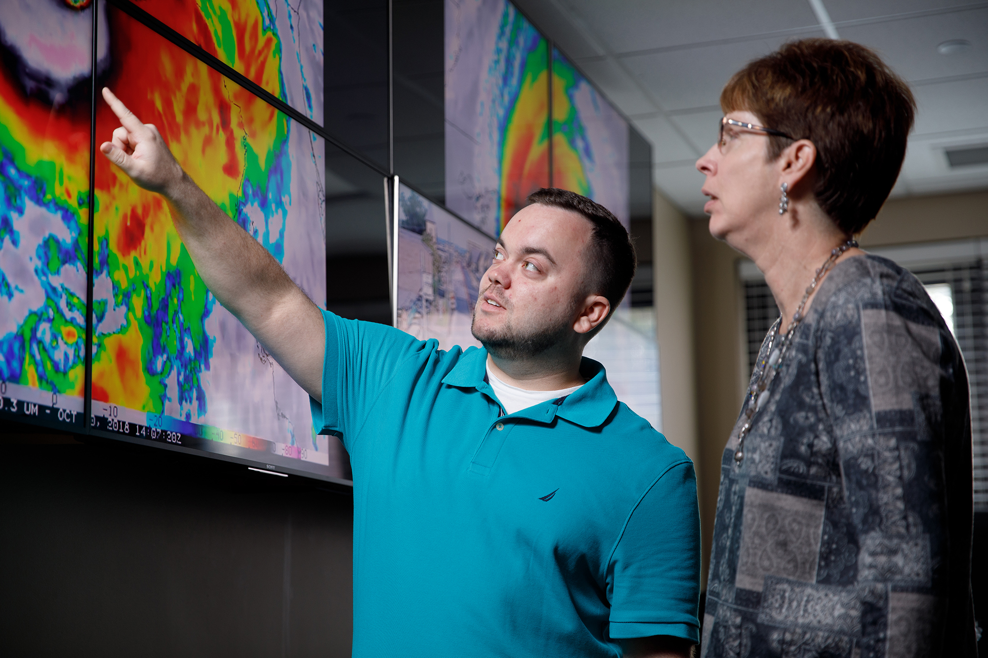 A meteorlogist points to a large screen displaying weather radar information while another scientist looks on.