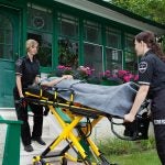 Two female paramedics escort a patient on a stretcher out of a home