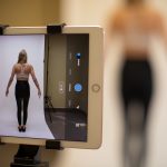 A woman in work-out clothes stands still while a tablet computer captures an image of her, shown on screen.