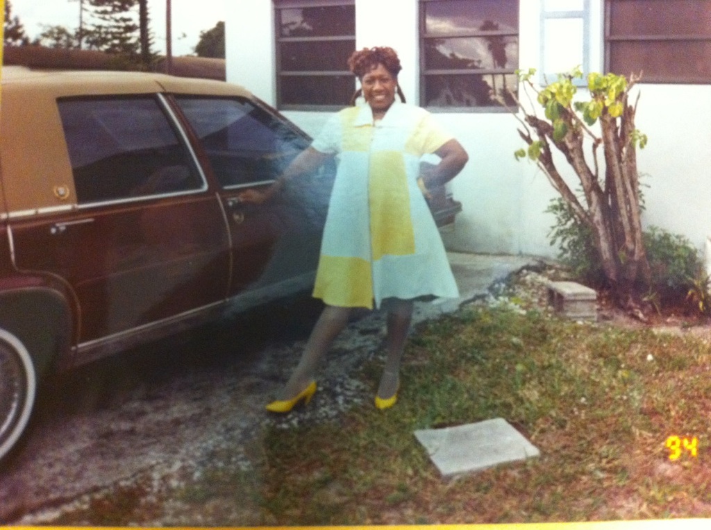 Green's mother in yellow dress and shoes in front of their home.