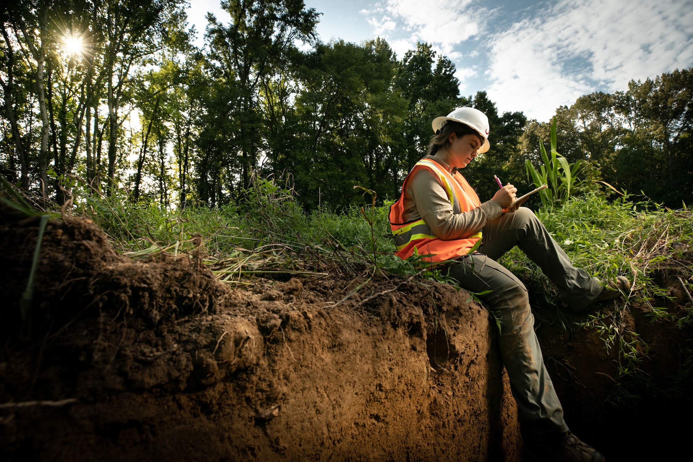 A researcher in safety gear sits on the edge of a hole in the forest taking notes.