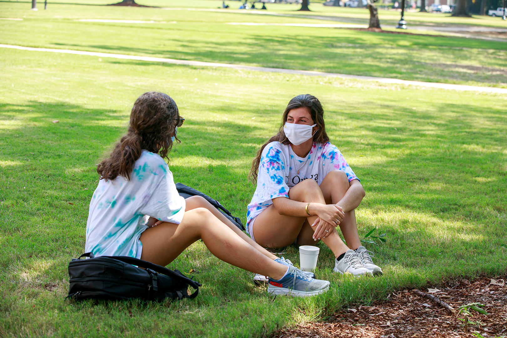 Two females with masks sit on the campus lawn.