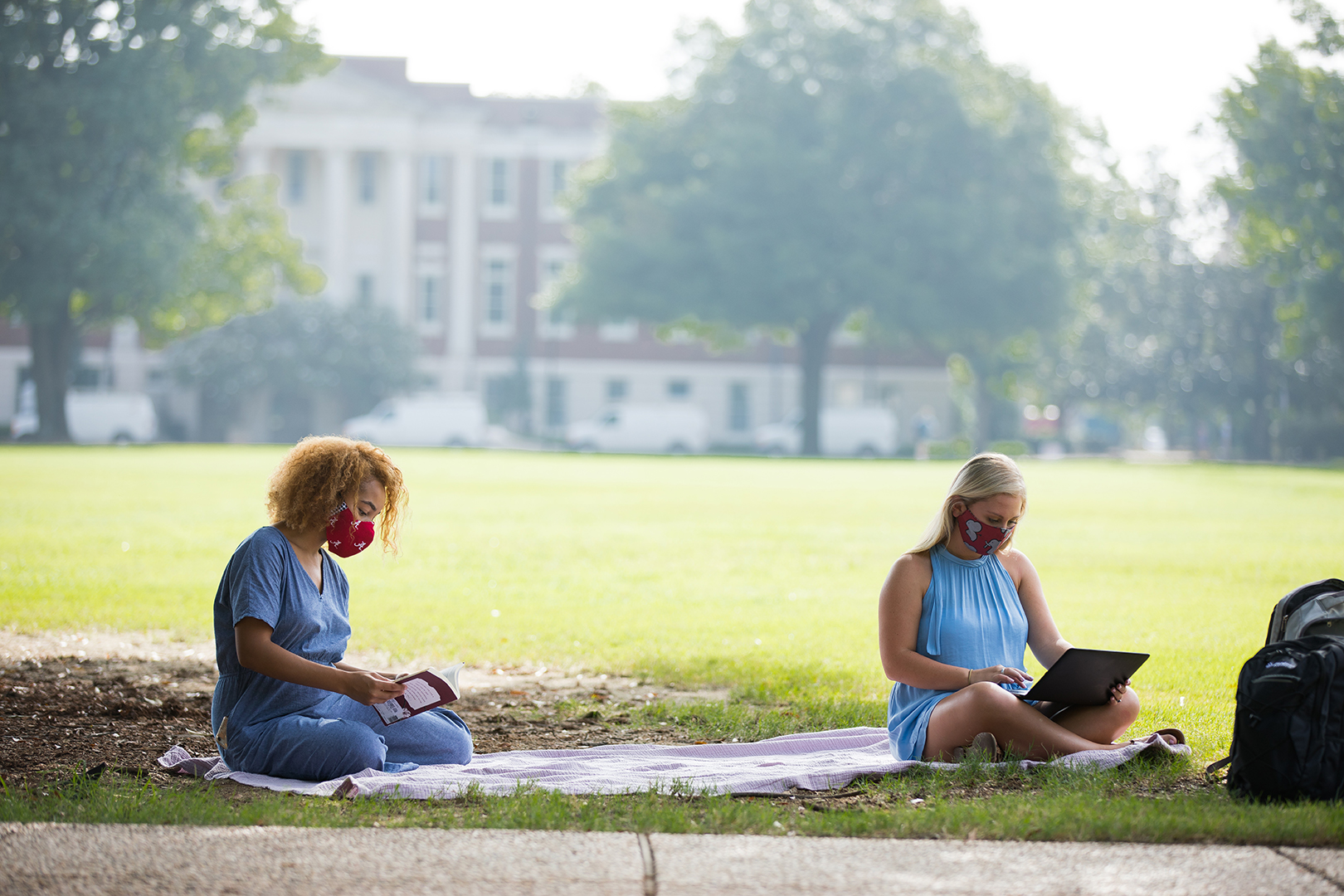 Two female students in masks sit on the UA campus lawn.