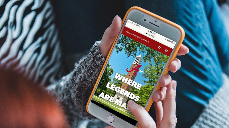 Hands hold a smartphone with the new ua.edu homepage on screen.
