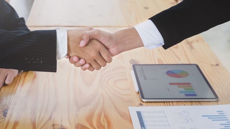 Two hands shake over a wooden table with business charts laying on top.