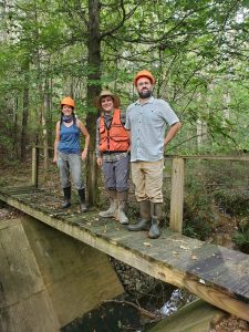 Three people stand on a wooden bridge over a creek ready for field research.