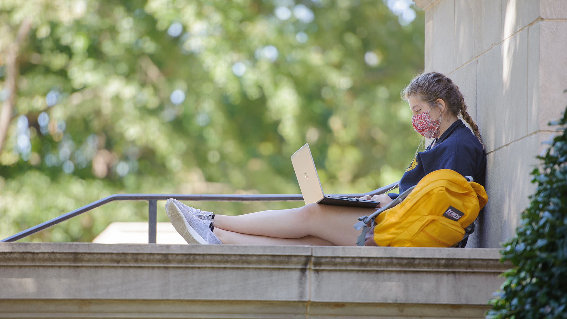 A female student wearing a mask works outside on her computer.