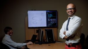 A man poses with arms folded in front a screen with engineering illustrations.