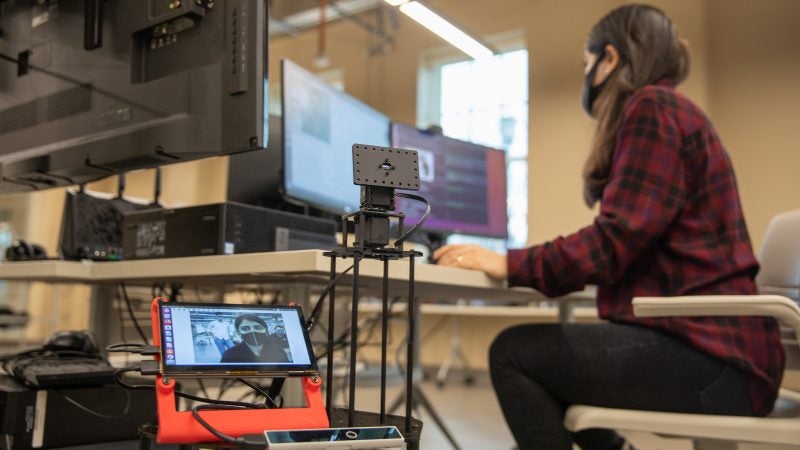 A woman in the background sits at a computer while a robot she controls is in the foreground displaying her face on a screen.
