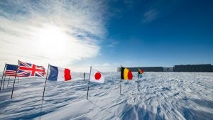 International flags fly around the South Pole on the ice with a research facility in the background.