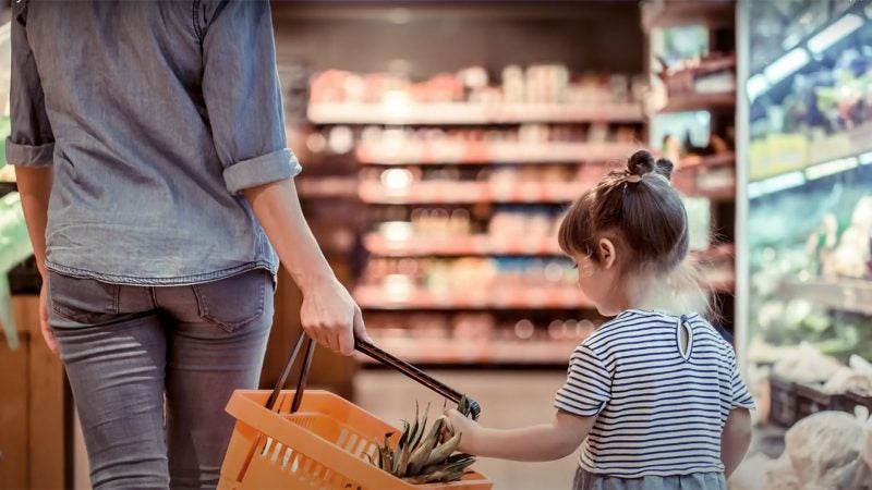 A woman and child carry a shopping basket through a grocery store.