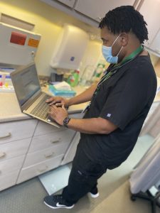 LaDArryl Banks wearing scrubs and a face mask types on a computer in the clinic where he works.