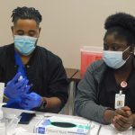 twp nurses in face masks examine a vial while sitting at a table