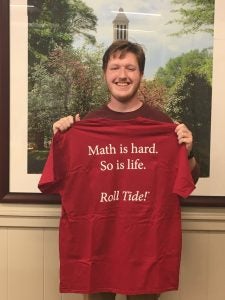 young man holds up a red t-shirt that reads, "Math is hard. So is life. Roll tide!"