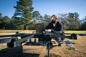 A man squats to type on a computer connected to a large drone in a field.