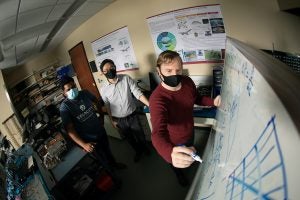 Three men with masks on gather around a white board with scientific schematics.