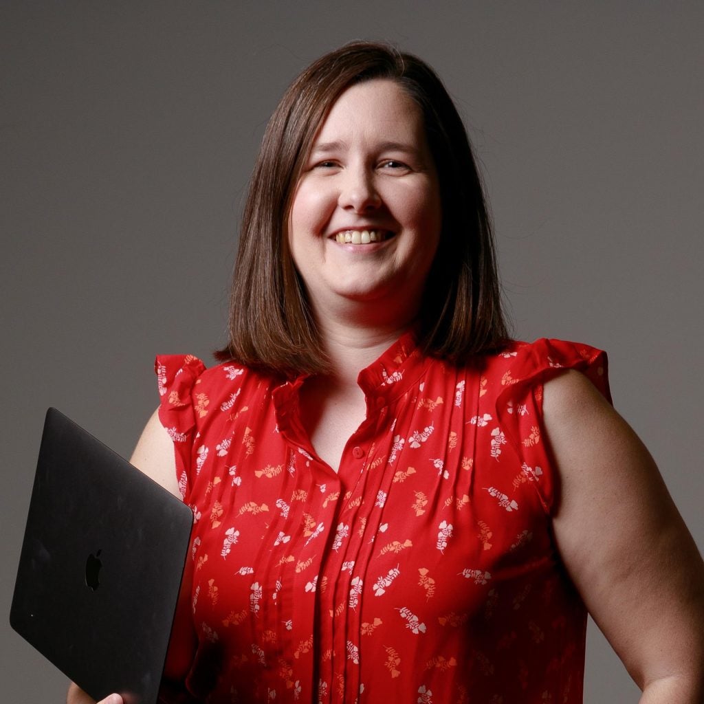 A woman in a red shirt with a small floral pattern, smiles while holding a laptop computer.