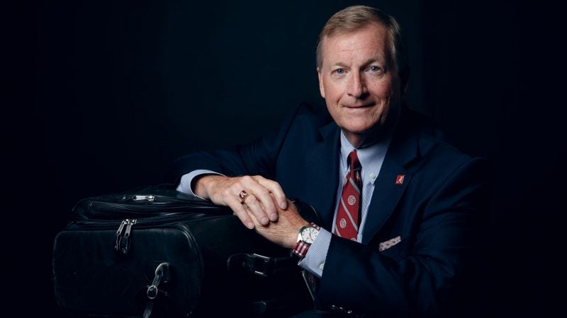 A man sits in front of a black background, leaning on a black suitcase with his hands crossed.