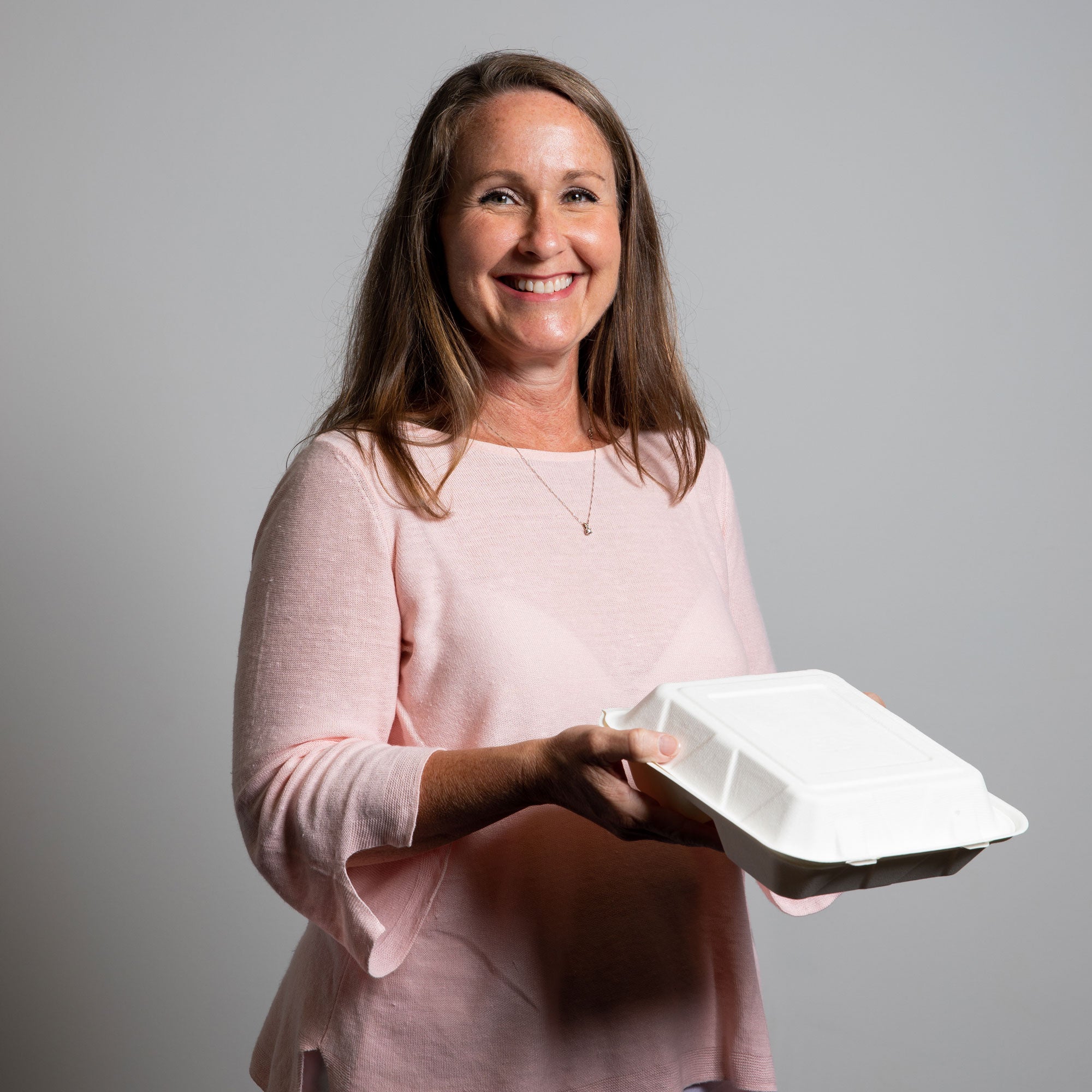 A woman in a light pink mid-length sleeved blouse is holding a to-go box while smiling.