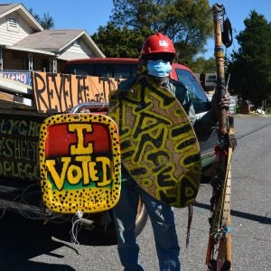 Artist Joe Minter stands next to a truck wearing some of his art, including a colorful sign that reads "I voted."