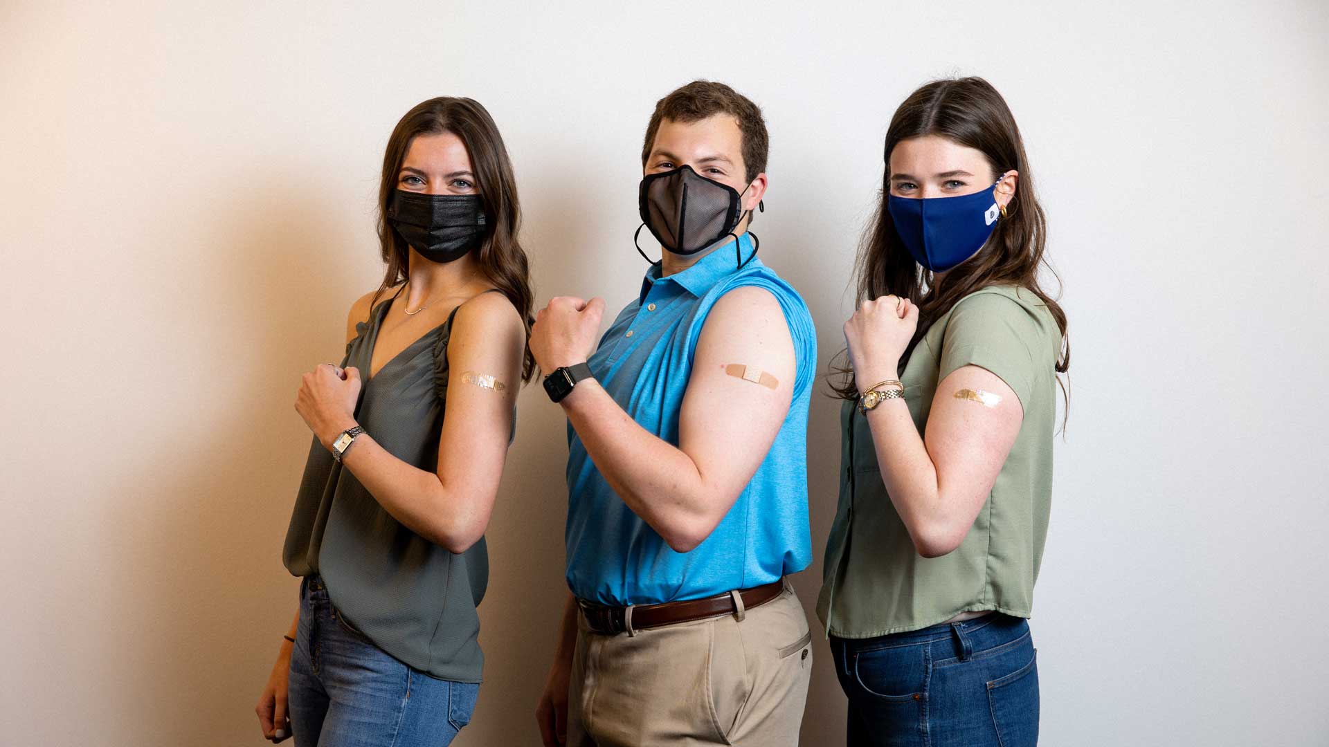 two female students and one male student stand wearing masks with their left arms flexing to show bandaids where they received the covid-19 vaccine
