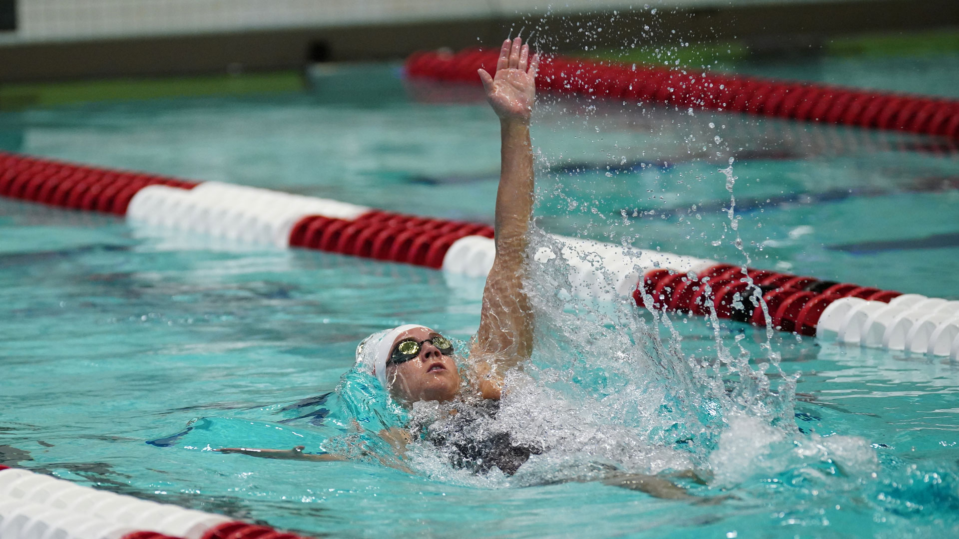 A woman is swimming the backstroke, with one arm getting ready to paddle. Crimson and white lane lines are also shown in the pool.