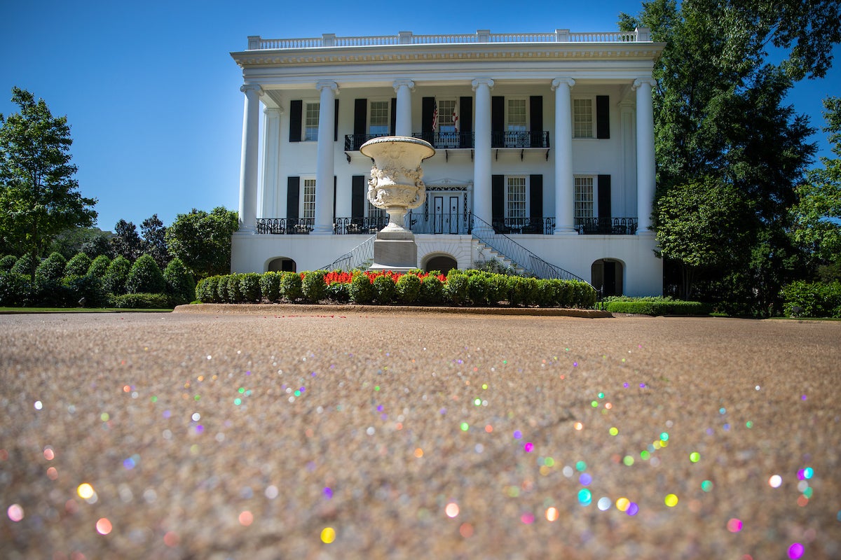 In the foreground, confetti of all colors is on the ground. A white Greek Rival styled house sits behind a statue of an urn.