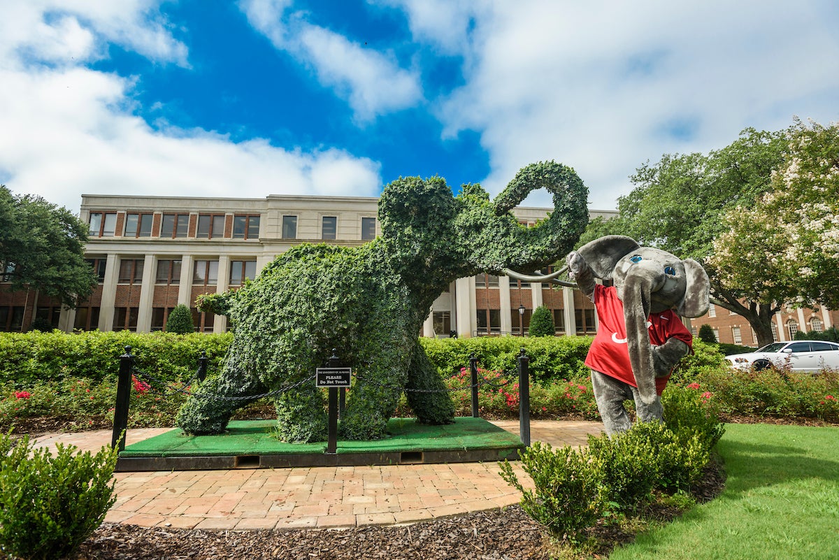 An elephant mascot stands next to a vibrant green elephant topiary.