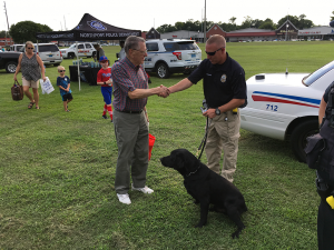 A man shakes the hand of a police officer at an outdoor event. A K 9 dog sits near him.