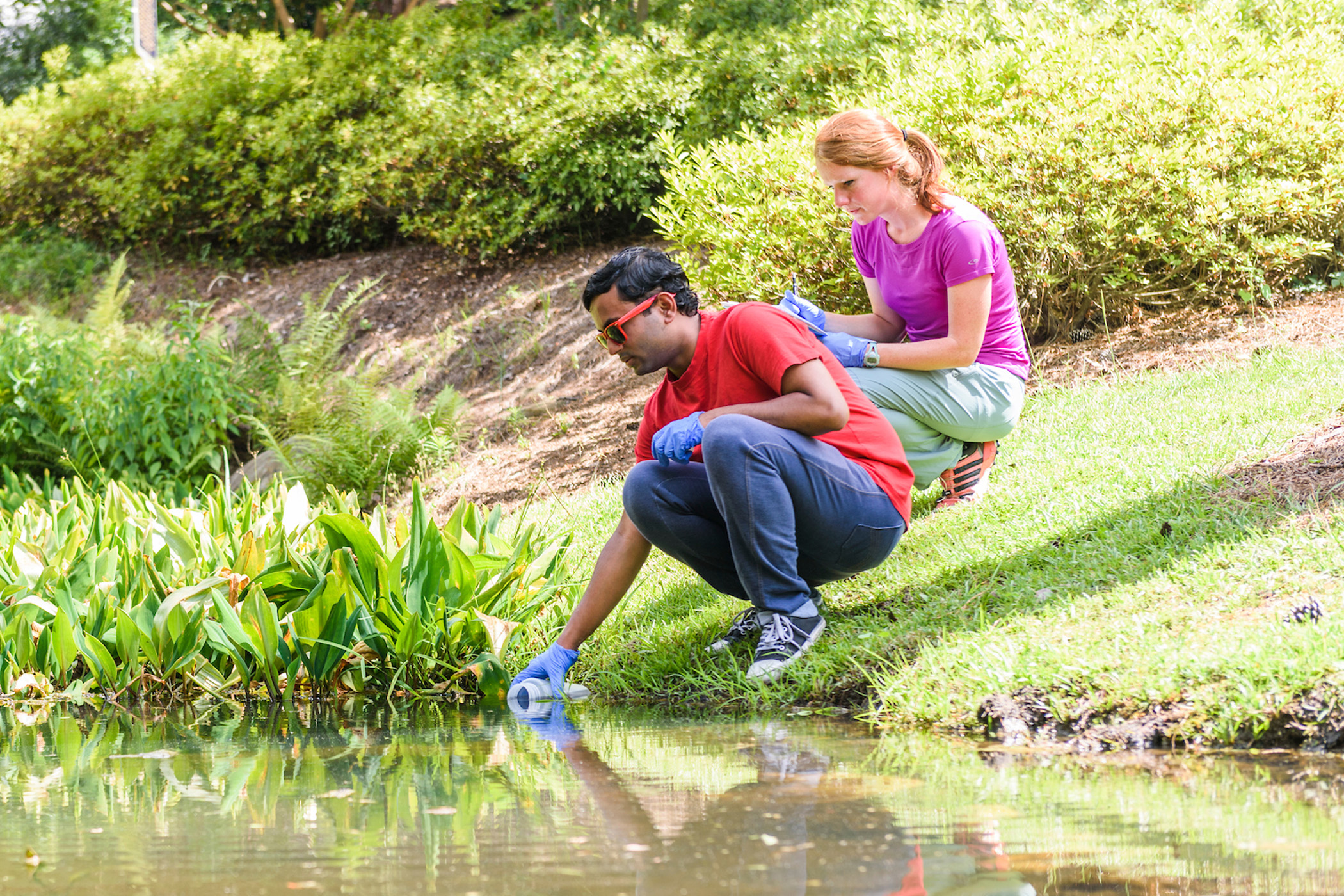 A student collecting a water sample from a pond while another student takes notes
