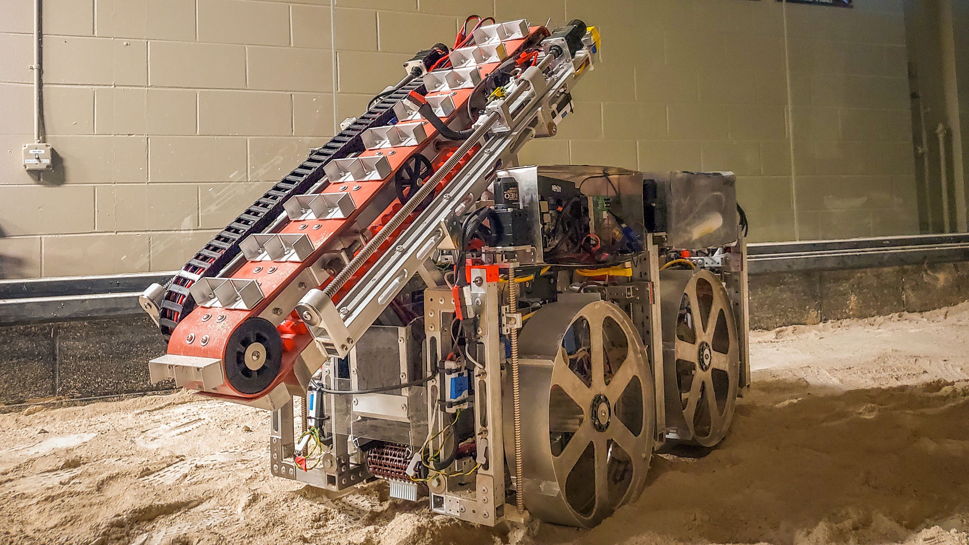A robot with four wheels and a conveyor belt for extraterrestrial digging against a wall that displays six NASA championship flags