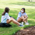 Two female students sit on the Quad's grass while wearing masks