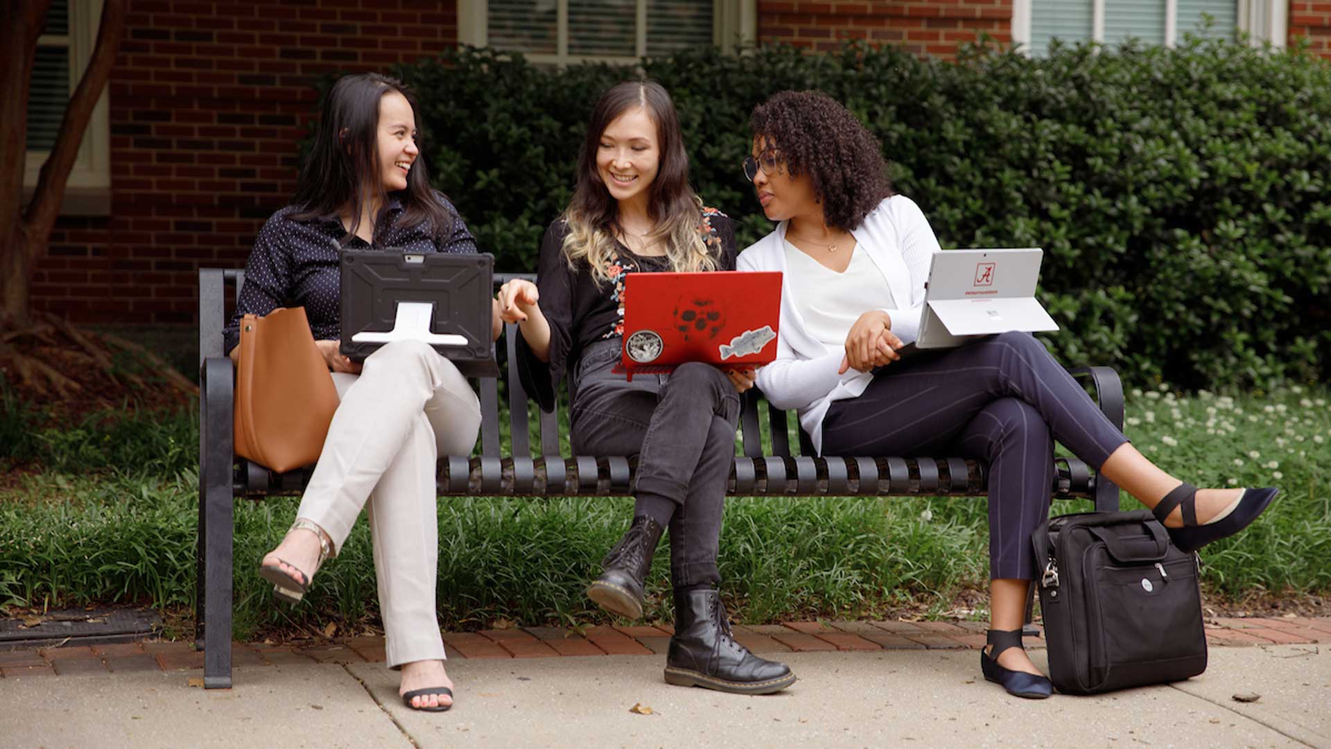 Three women sitting on a black bench outside are chatting. Each woman has a laptop.