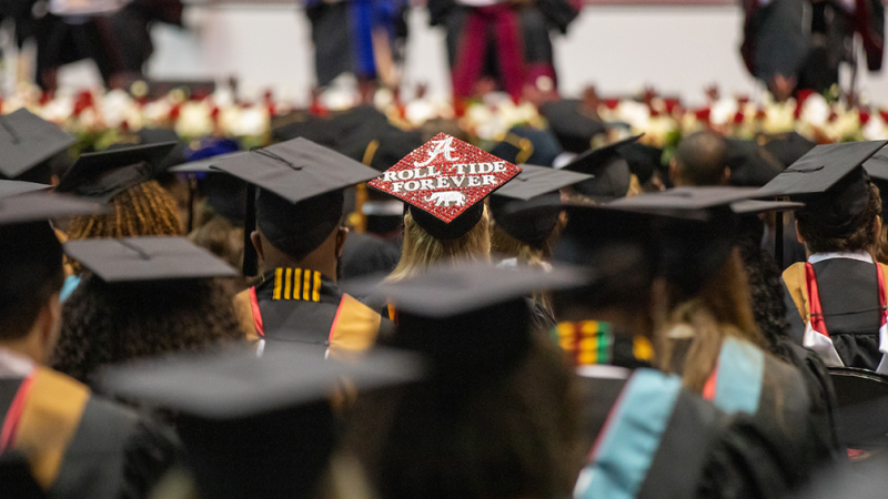 Students wearing mortar boards at a commencement ceremony. One mortar board says Roll Tide always.