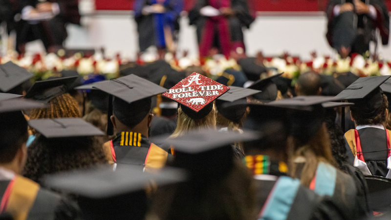 Students wearing mortar boards at a commencement ceremony. One mortar board says Roll Tide always.
