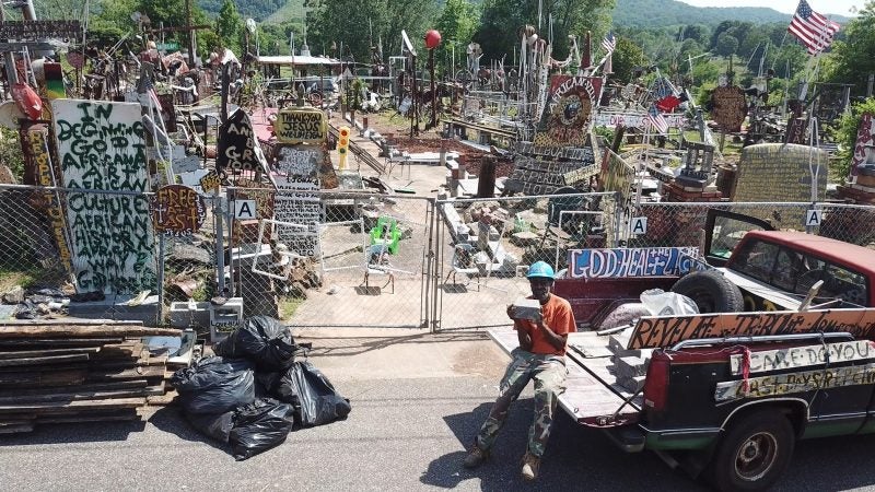 A man on the tailgate of a pickup truck sits before his yard of scultpures.