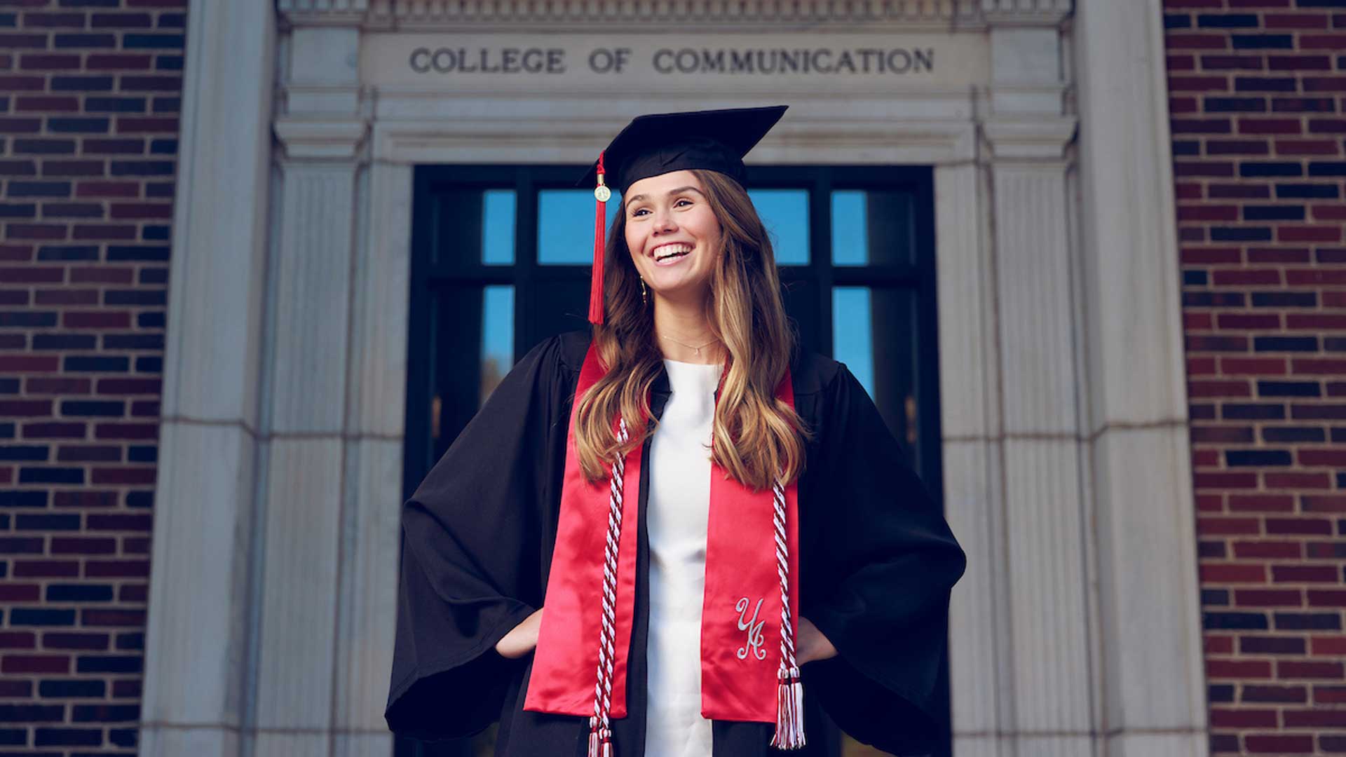 A girl with light to medium brown hair poses next to a concrete pillar while dressed in a cap and gown.