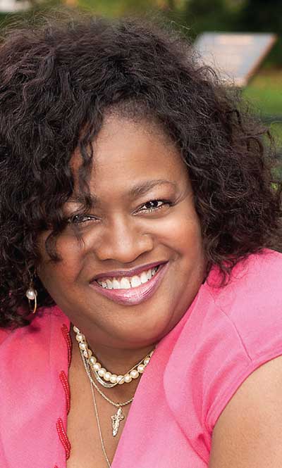 A Black woman smiles at the camera. She is wearing a pink shirt, with a pearl necklace.