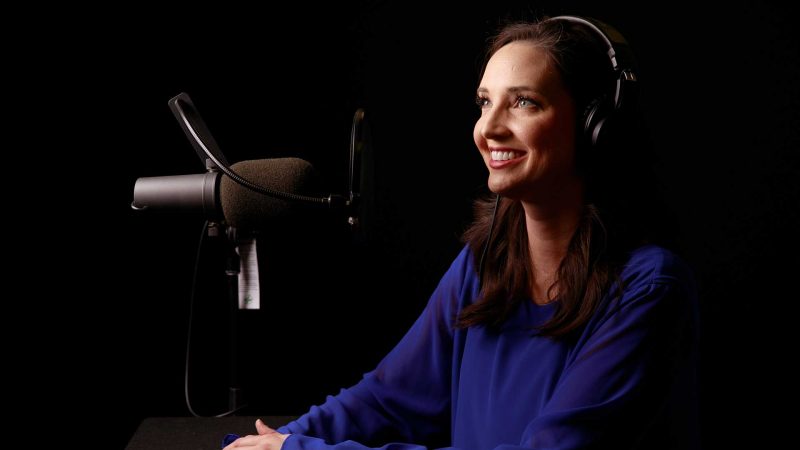 A woman with brown hair sits in front of a black background, smiling off camera. She is wearing a blue shirt and has a microphone next to her.