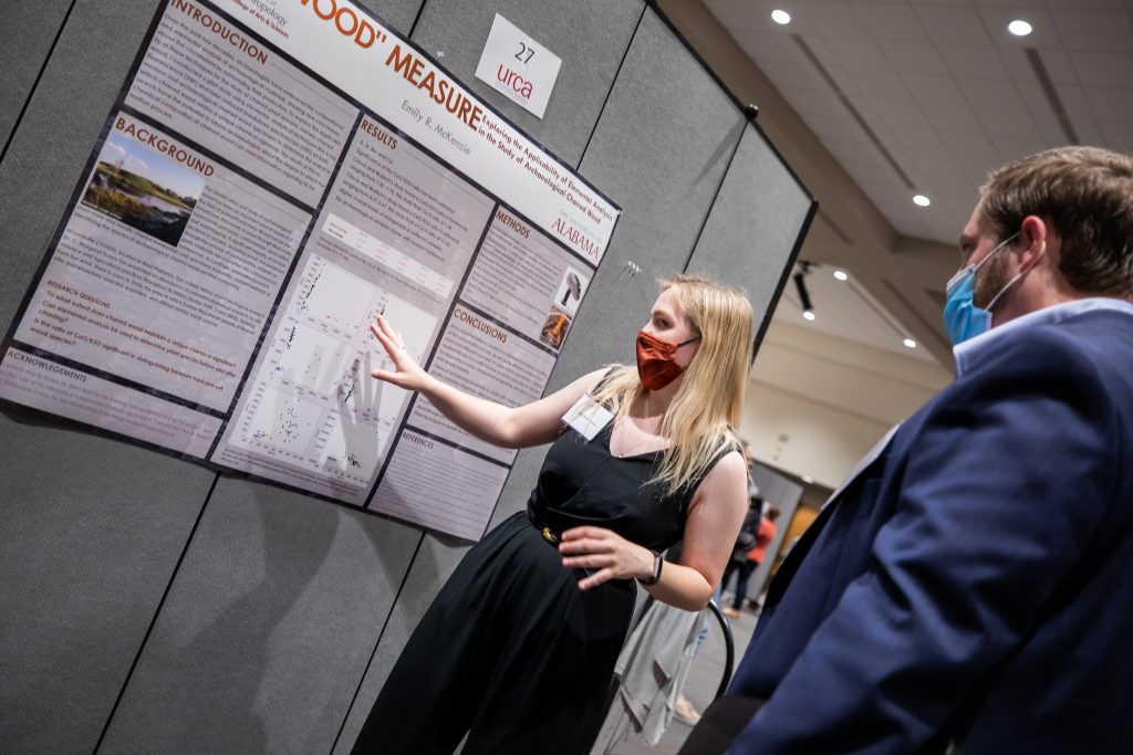a female student points to a research poster mounted to a wall