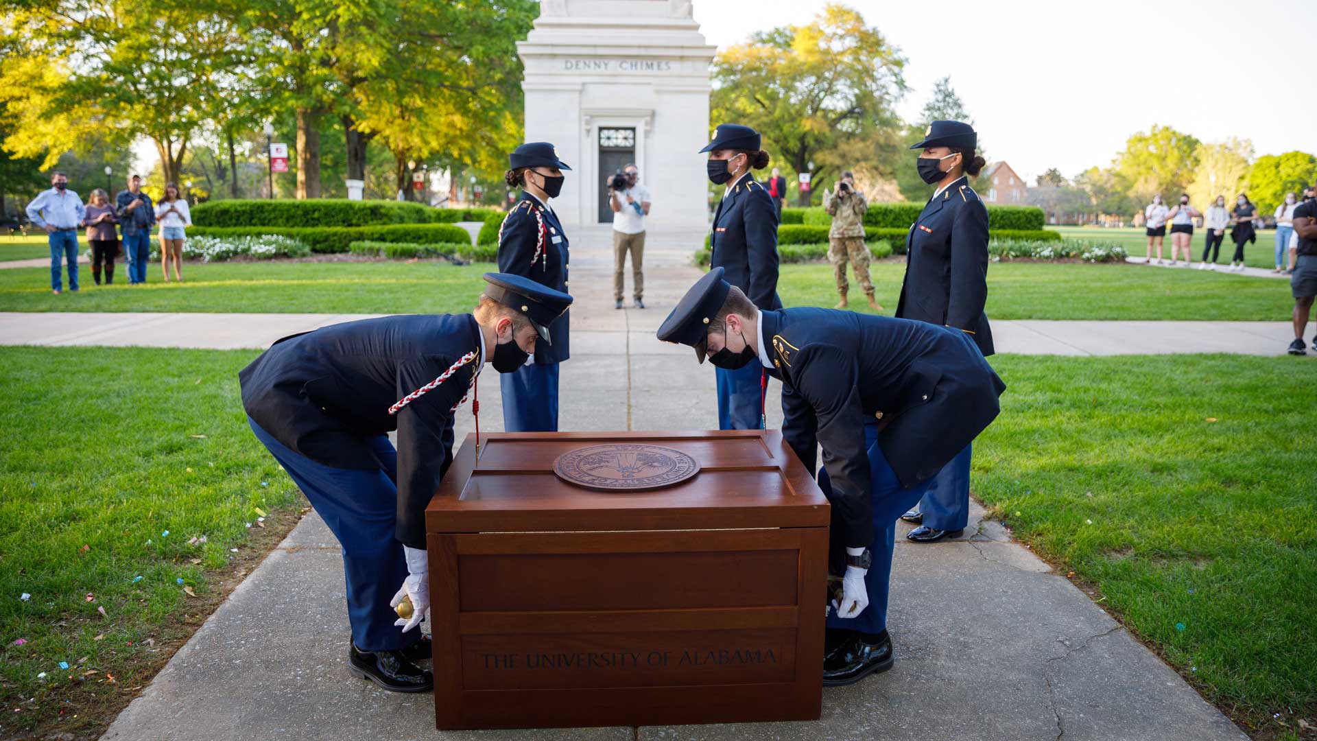 two young men in uniforms lift a wooden chest with the Univerisity of Alabama seal on top of it
