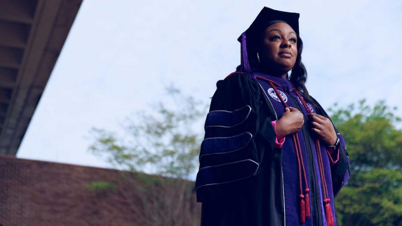 A Black female student looks off camera while dressed in her cap and gown. She will earn her JD this spring.
