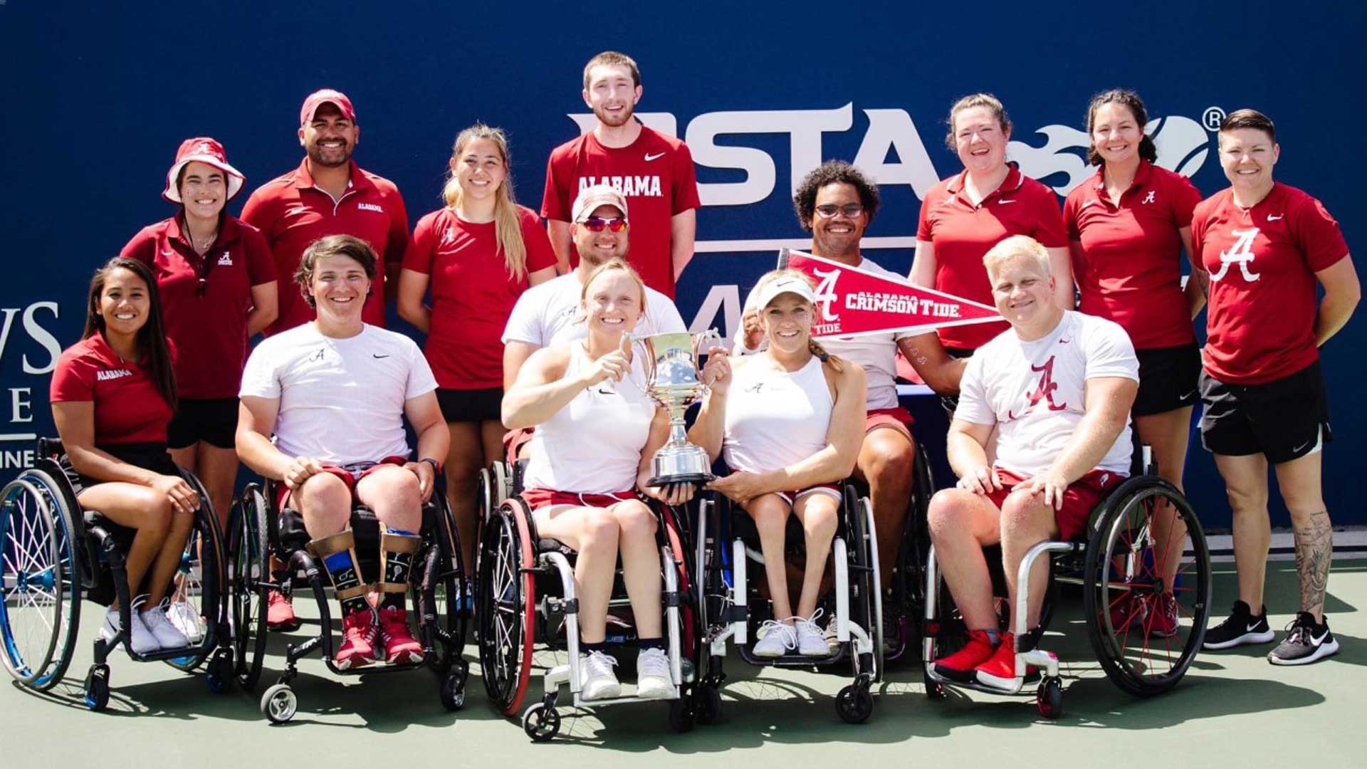 On a dark navy background, 13 people are posing for the camera. Four of the subjects are in white shirts and sitting in wheelchairs.