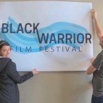 Two students, one male and one female, hold up a poster that says "Black Warrior Film Festival"