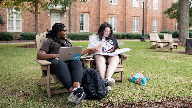 two young women study in chairs on the Quad with books and laptops