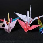 Six paper cranes are folded and sitting on a black background. One crane is coral pink, one is lavender, one is tan, two are multicolored and one is folded on Hiragana practice paper.