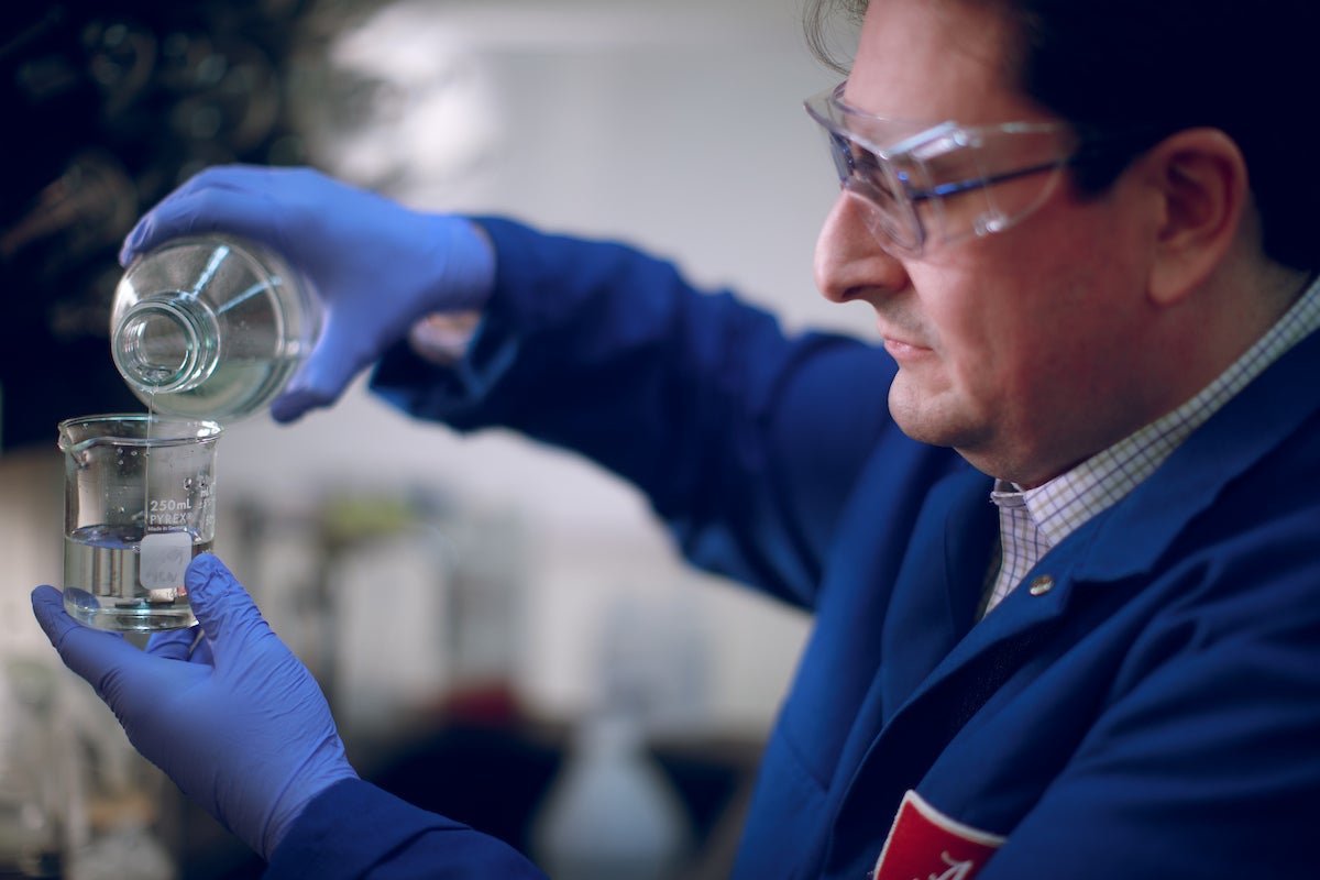 A man in safety goggles and blue lab coat pours water into a container in a lab at The University of Alabama.