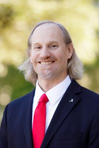 A professional headshot outside of a man in long hair in a dark suit and red tie.